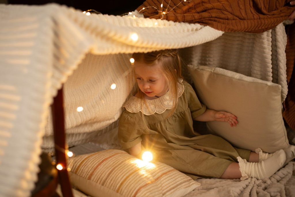 Petite fille assise dans une cabane de couvertures éclairée par une guirlande lumineuse, ambiance douce de soirée pyjama